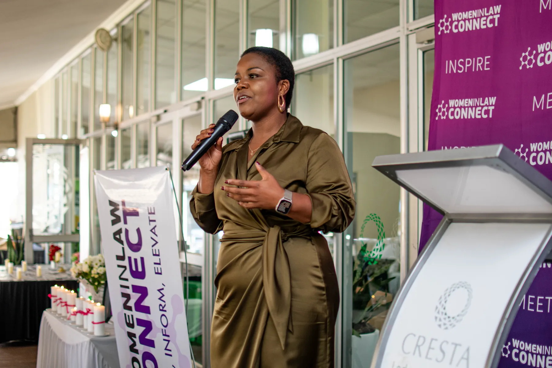 A woman in an olive green satin wrap dress speaks confidently into a microphone. She stands between a white "Women in Law Connect" banner and a purple backdrop featuring the words "Inspire" and "Meet."