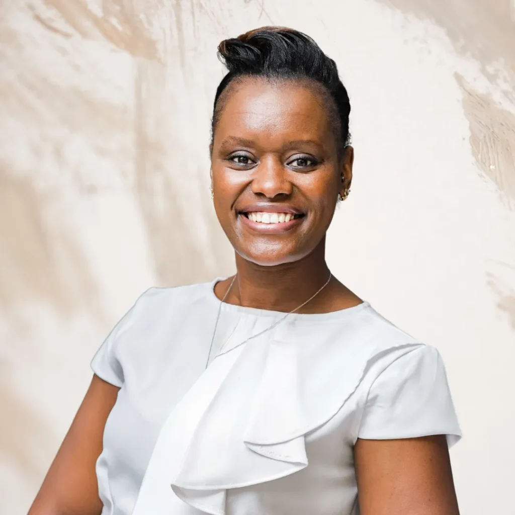 A professional head-and-shoulders portrait shows a Black woman with a confident smile, looking directly at the camera. She has short, dark hair styled in a textured, upward-swept look and is wearing a light grey or white short-sleeved top with an elegant ruffled detail down the front. The background is a soft, neutral-toned abstract texture with subtle beige and white brushstroke-like patterns.