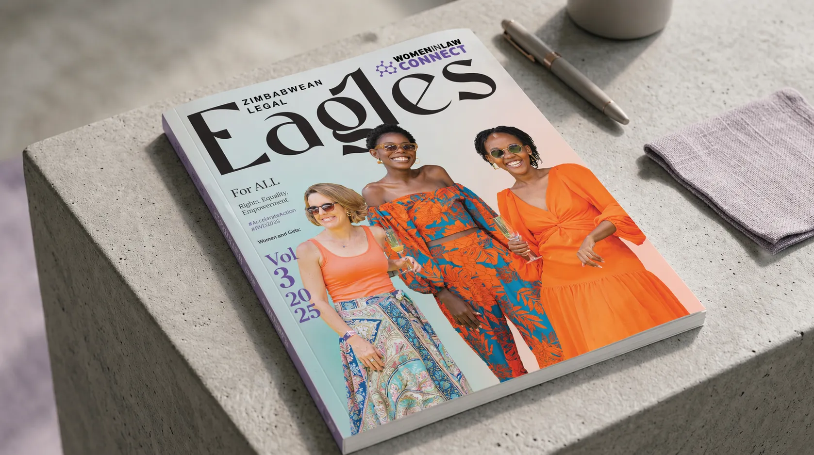 A professional mockup features a physical copy of Legal Eagles Magazine resting on a sleek, dark wooden desk. The magazine cover displays three women in vibrant orange and teal outfits smiling together against a pastel background.